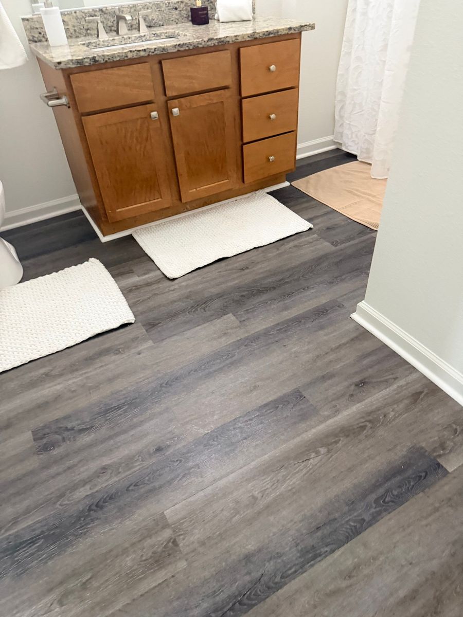 Freshly cleaned apartment bathroom with a granite-topped wood vanity, spotless gray plank flooring, and white bath mats.