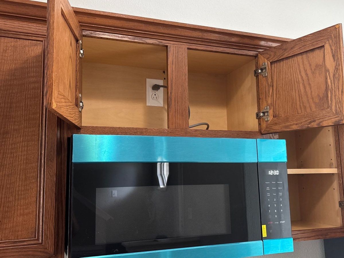 Newly installed over-the-range microwave, protective film still on, mounted beneath open oak cabinets in an apartment kitchen.