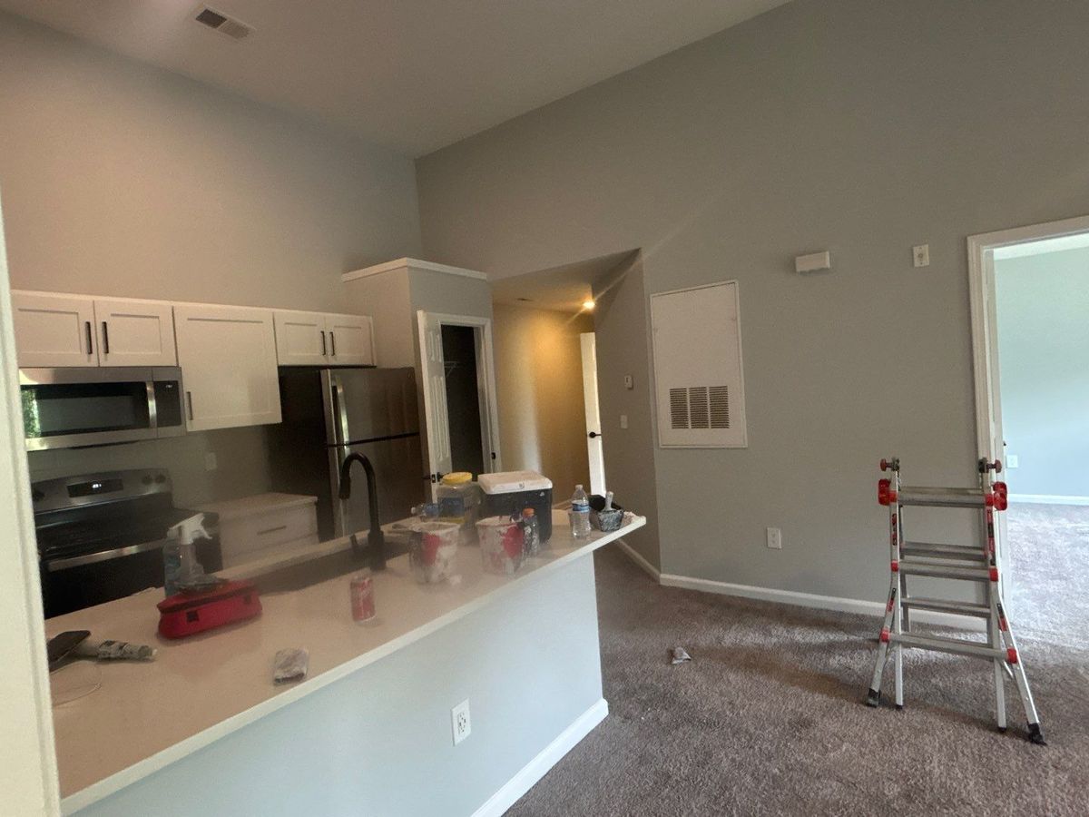 Apartment living room and kitchen mid-repaint: freshly painted gray walls and white trim, with a step ladder and paint supplies on the counter.